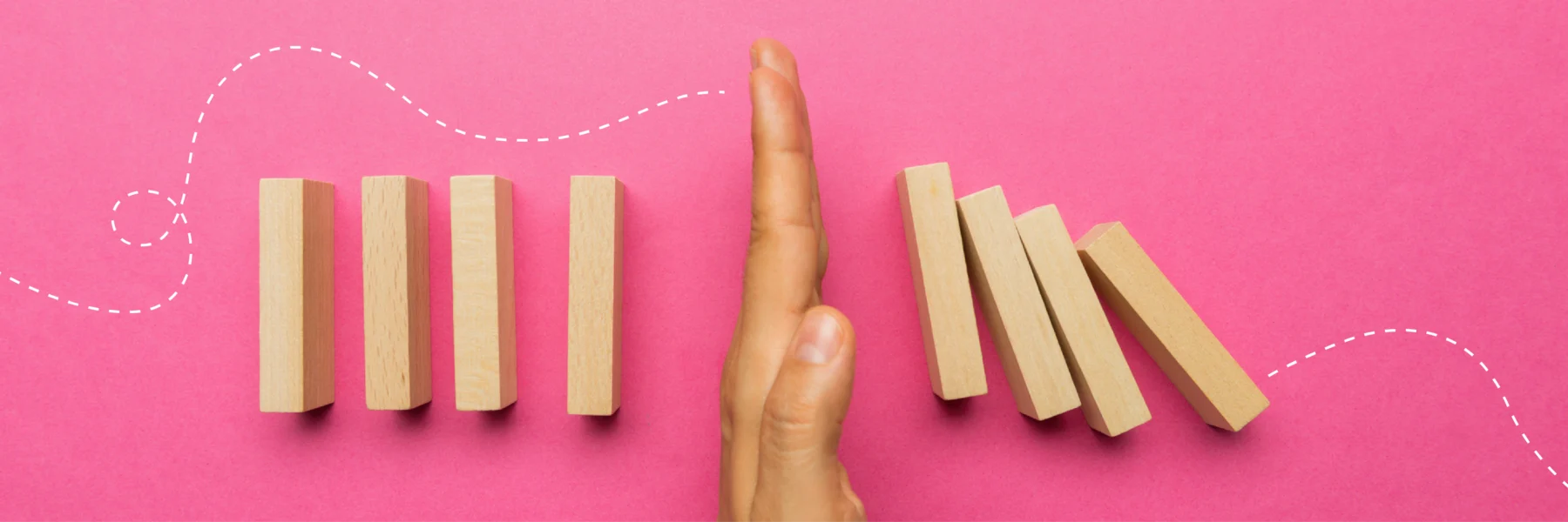 A hand stops a row of wooden dominoes from falling on a pink background with a dashed line.
