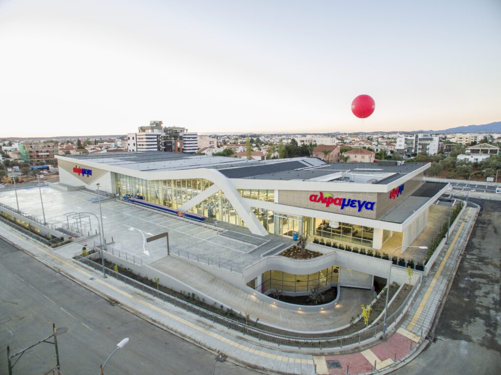 A large, modern supermarket building with "Alphamega" signage, surrounded by empty parking lots and city buildings in the background.