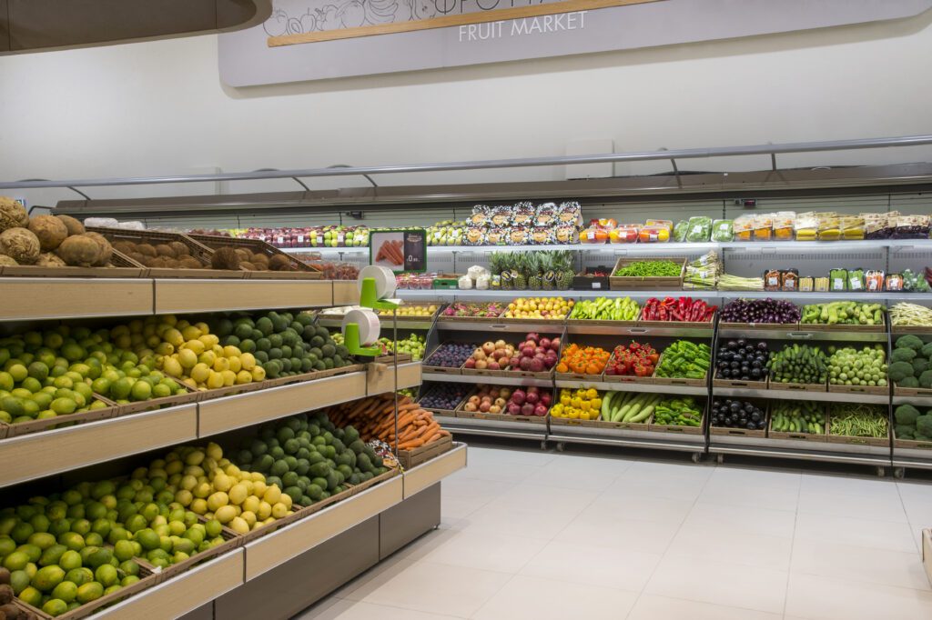 A grocery store produce section displays neatly arranged fruits and vegetables on shelves and in bins, including citrus, apples, carrots, and leafy greens.