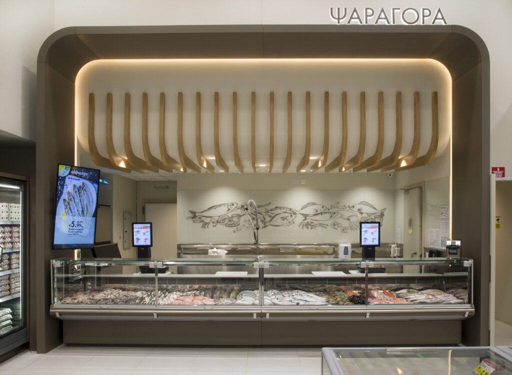 A modern seafood counter with various fish and seafood on display, illuminated by overhead lighting, and a decorative fish mural on the wall behind. The word “ЧАРАГОРА” appears above.