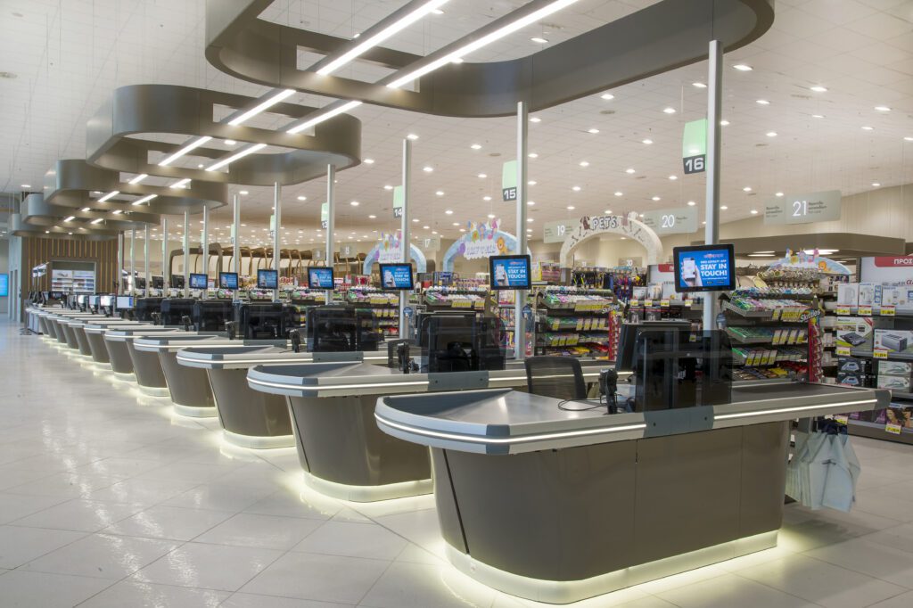 A row of empty, modern supermarket checkout counters with digital displays, under bright overhead lighting in a spacious retail environment.