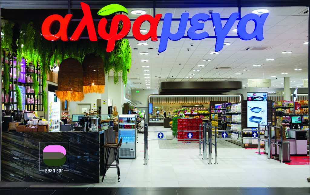 Front entrance of an Alphamega supermarket with a coffee bar on the left, visible signage, bright lighting, and aisles of products in the background.