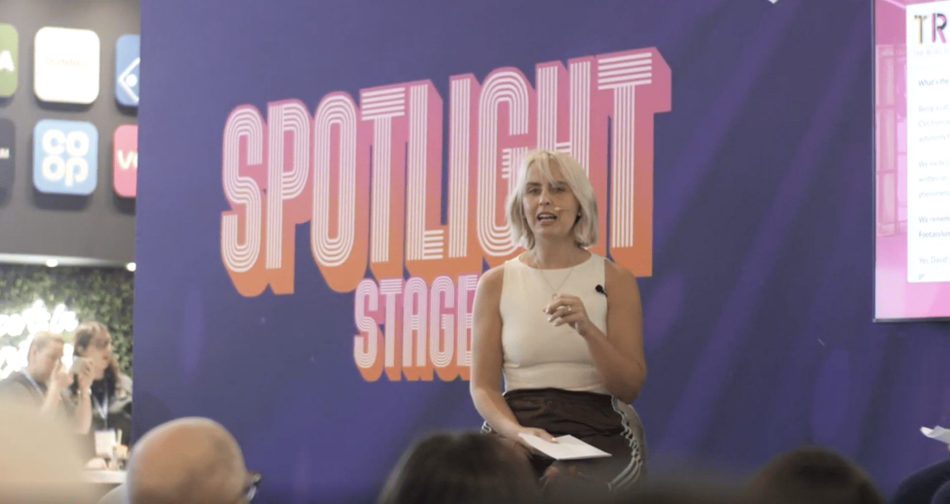 A woman with short blonde hair speaks on stage in front of a large "Spotlight Stage" sign, holding papers and wearing a microphone headset. Audience members are visible in the foreground.