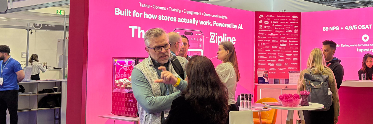 A man speaks to a woman at a trade show booth with bright pink branding for Zipline. Several people interact around the booth, which displays promotional materials and digital screens.