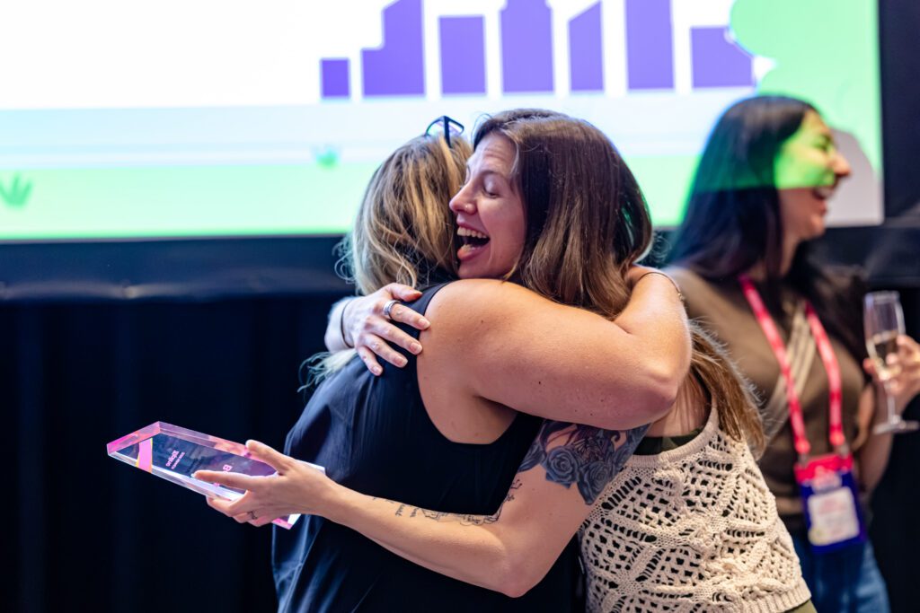 Take a Bow: Celebrating Zipline’s 2026 Summer Camp Award Winners 1 Two women embrace in celebration, one holding a transparent award plaque. Another woman in the background holds a drink and smiles. A presentation screen is visible behind them.