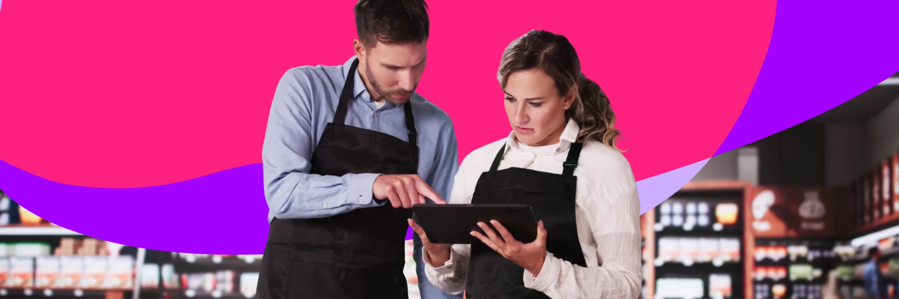 Two people wearing black aprons stand in a store aisle, looking at and discussing information on a tablet. Bright abstract shapes fill the background above store shelves.
