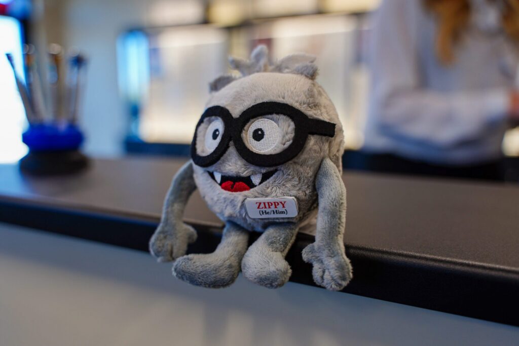Zippy in Action: Four Real-World Use Cases for Zipline's Frontline AI Assistant 3 A gray plush toy with black glasses and a name tag reading "Zippy (the Ultium)" sits on a counter; a person stands blurred in the background.