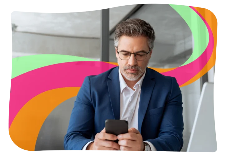 A man in a blue suit and glasses looks at his phone, sitting indoors with colorful abstract shapes in the background.