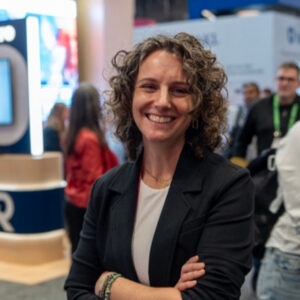 A person with curly hair and a blazer stands smiling with arms crossed at an indoor event with people and booths in the background.