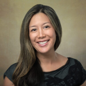 A woman with long brown hair and a black top smiles in front of a softly textured, neutral-toned background.