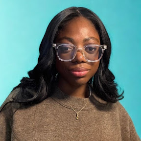 Woman with long black hair and clear glasses wearing a brown sweater and gold necklace, standing in front of a solid teal background.