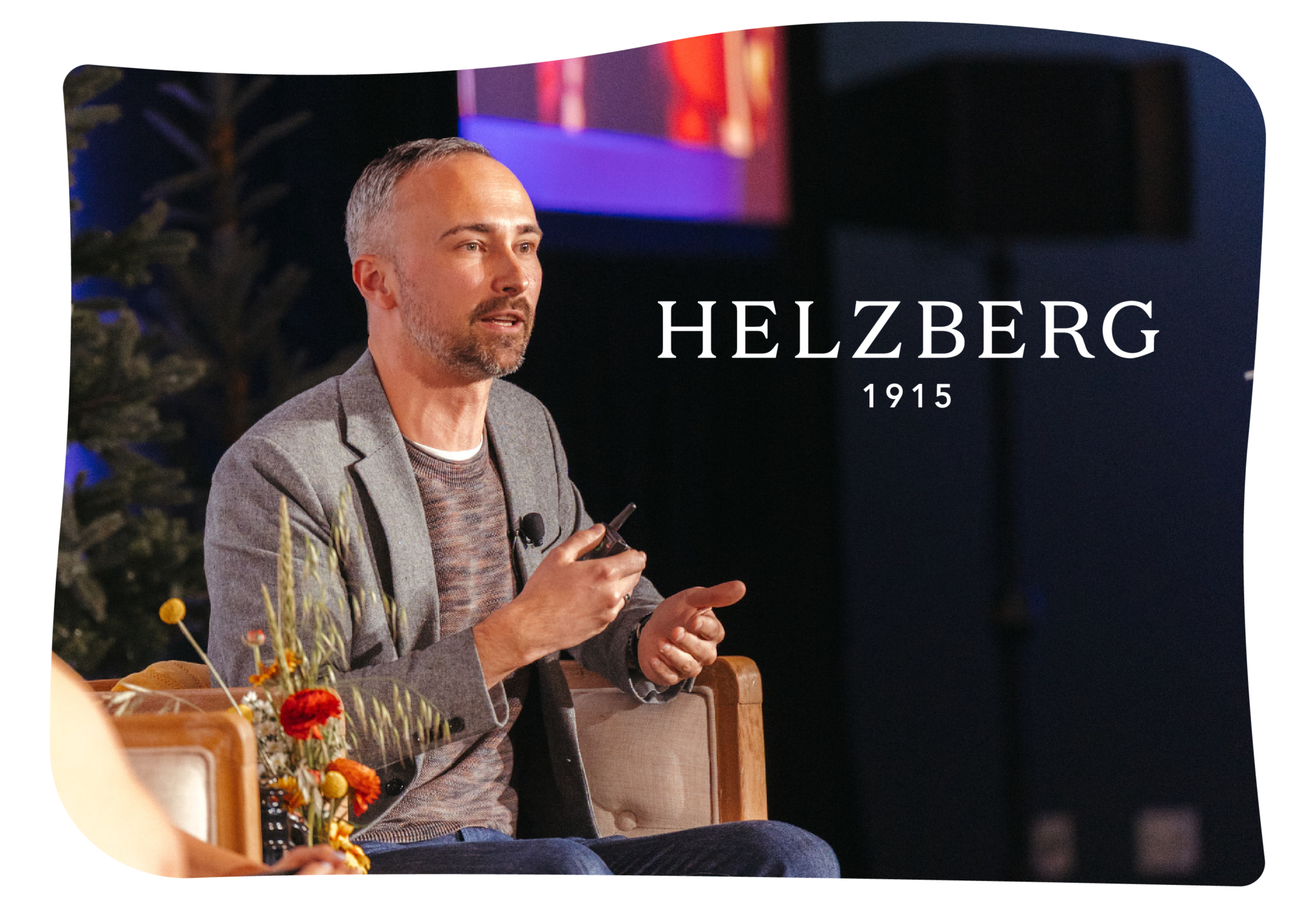 A man with short gray hair and beard speaks while seated on stage, holding a pen. The background displays the text “HELZBERG 1915.”.