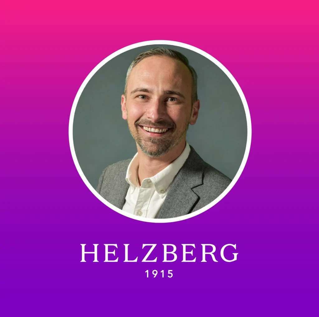 Smiling man with short hair and beard in a gray blazer and white shirt, inside a white circle on a pink to purple gradient background with text “HELZBERG 1915” below.